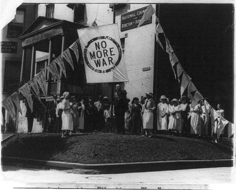 Black and white photograph showing a large group, mostly women, in front the National Council for Reduction of Armaments headquarters, in the same building as the National League of Women Voters headquarters in Washington, D.C. Thy arre raising a large white banner which says 'No more war'.