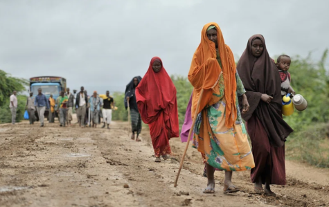 Refugees in Africa are driven out of their village. Many people walk away from a bus in the distance towards the camera. In the foreground there are 3 women one carrying a child wearing brightly coloured clothing.