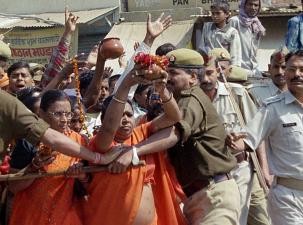 Two older Indian women wearing orange are in a crowd being held back by male Indian law enforcement wearing green uniforms
