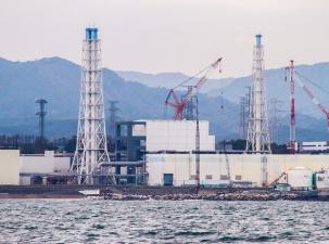 White nuclear power plant with two red and white cranes | Choppy dark blue water in the foreground and hazy mountains in the background