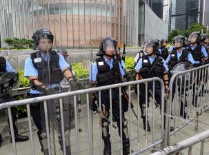 5 police with helmets and shields behind a metal barrier infront of a glass building in Hong Kong