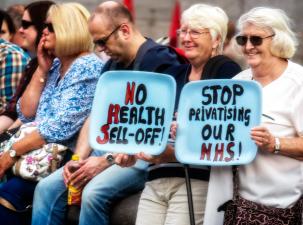 A group of older men and women sitting on chairs smiling holding painted plates about not selling the NHS