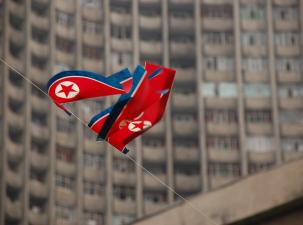 Two red and blue North Korea flags flying in the wind on a string with a block of grey buildings in the background