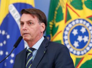 Serious looking white man with blue eyes wearing a suit speaking on a stage infront of the Brazilian flag