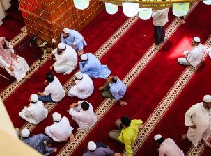 Many male muslim worshippers sit on the red carpeted floor of a mosque wearing tradional white skullcaps