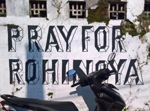 Pray for Rohingya mural painted in black on a white crumbling wall - black scooter parked infront