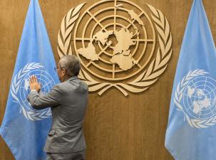Gold UN logo on a wooden wall sits between too large blue UN flags, a dark skinned older man with greying hair wearing a grey suit stands and straightens one o the flags