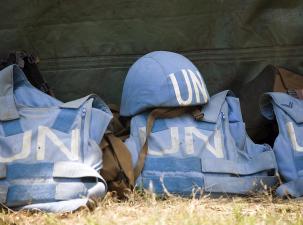Three light blue UN vests laying on the grass - UN written in white in big letters in the middle - one UN helmet sits on top