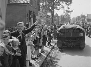 Black and white image of an army truck driving down a road lined with waving children
