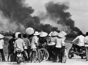 Black and white image of two huge smoke plumes and many Vietname onlookers watching the smoke wearing traditional cone shaped hats