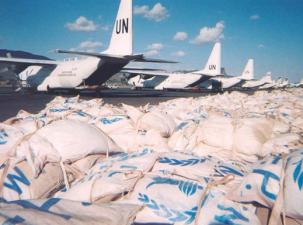 White sacks of food laying all over an airport runway with the blue UN logo printed on them - 7 white UN planes lined up in the background under a blue sky
