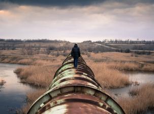 Dark image of a man walking into the distance along a huge rusted gas pipeline which goes through wetlands with dry grass growing through the water