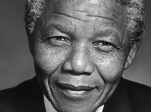 A black and white photograph of Nelson Mandela wearing a black suit and leaning into the camera