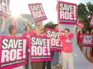 A group of people wearing pink tshirts stand holding posters saying Save Roe!