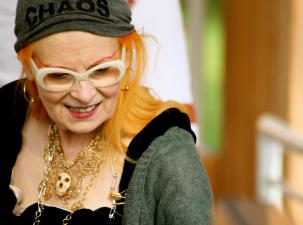 Cultural icon Vivienne Westwood sports her trademark orange hair at an Active Resistance to Propaganda event. She is smiling, looking away from the camera, wearing an assortment of necklaces and a grey headband that reads 'chaos'