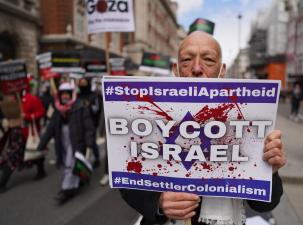 An older white gentleman stands in the street holding a large white sign which says ' Boycott Israel' the man is part of a larger protest which we can see part of in the background