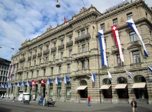 The Crédit Suisse headquarters in Zürich built between 1873-1876. The four storey building is sand coloured, has many flags outside, and has 2 statues above the main entrance.