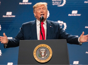 Image of former U.S. President Donald Trump standing behind a podium infront of a blue background. He has his arms wide open and is talking to the crowd 