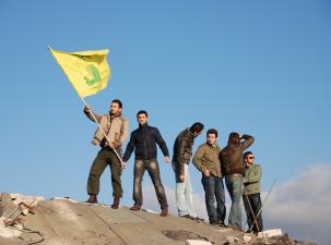 Six you men stand on top of a destroyed building, one of them is holding the yellow fllag of Hezbollah