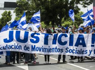 Protesters in Nicaragua demand justice. They march down a street holding a large white banner that says ' Justicia' in blue. Many of the protesters hold Nicaragua flags.
