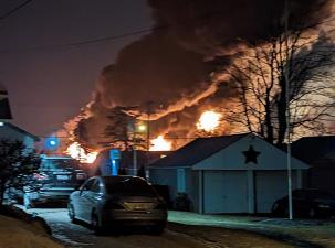 Photo taken overlooking the 2023 Ohio train derailment during the night. There is a large plum of dark smoke billowing towards the sky, 3 fires can be seen in the background.