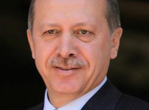 Portrait of the Turkish President Erdoğan. He wears a black suit with a blue striped tie and a small pin of the red Turksih flag on his lapel.