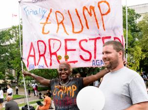 People celebrate outside the courthouse as Trump arrives for his arraignment. A woman wearing a black tshirt that says 'Trump arrested' is holding a large white banner which reads the same.