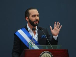 Photo of Nayib Bukele holding a ribbon of the flag of El Salvador, on a podium waving to the public.