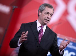 Nigel Farage speaking at the 2015 Conservative Political Action Conference. He stands infront of a red background wearing a black suit holding his hands open and looking away from the camera