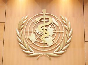 Gold logo of the World Health Organization hangs on a wooden wall. A snake is coiled around a staff infront of a mp of the world with the leaves of the UN symbol on both sides