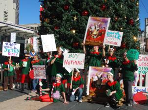 Protesters dressed as elves stand in the street holding signs which say 'Buy nothing' and 'Create don't consume'.