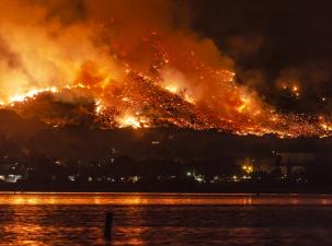 Image of a forest burning looking from across a lake. The sky is glowing orange, there are a lot of Embers in the air and smoke fills the sky.