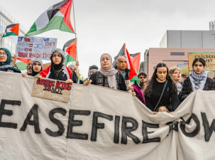 Protesters stand in the street calling for an end to the fighting in Gaza. They hold a large white banner which reads 'Ceasefire Now'