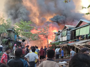MAF bombing Kyauktaw Township in Myanmar as part of their civil war. A large explosion and cloud of smoke fill the sky at the end of a street of wodden houses. Many onlookers standby. 