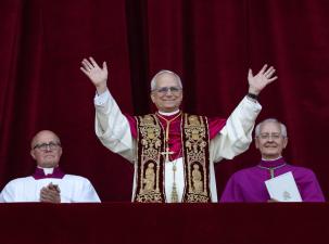 Pope Leo XIV addresses an audience with open hands, he is wearing white and purple robes and is smiling warmly.