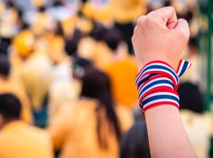 Adult hand wraps Thailand flag on a blurred protest in the background.