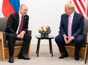 Trump and Putin sit at a table talking and smiling. They both wear suits and they have their countries flags behind them.