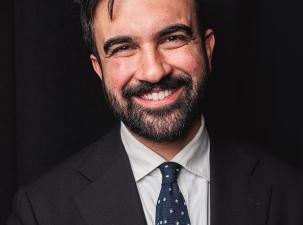 Zohran Mamdani wears a black suit and white shirt and smiles warmly at the camera infront of a black background.