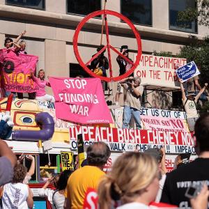 Colourful image of a bus decorated with peace symbols there are protesters riding on top displaying anti-war signs and against funding the war
