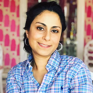 A smiling woman with dark hair styled in loose waves, wearing a plaid shirt, seated in a brightly decorated caf&eacute; with pink accents.