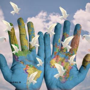 Two open palms are painted with a map of the world, in the background there are white doves in the air in front of a blue cloudy sky