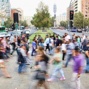 Colourful city street scene with many people rushing to cross a busy road