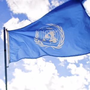 Light blue flag flying in the wind with the white UN logo in the centre | Blue sky and white clouds in the background