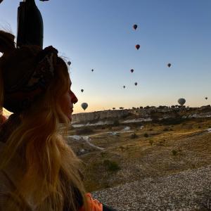 A silhouette of a women from a hot air balloon looks out into the sunrise. There are many other balloons in the sky in the background. 
