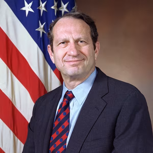 A professional headshot of a man in a suit, smiling, standing in front of an American flag backdrop.