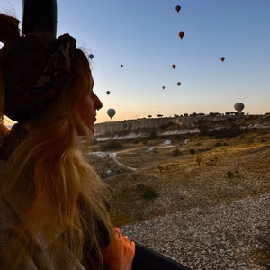 A silhouette of a women from a hot air balloon looks out into the sunrise. There are many other balloons in the sky in the background. 