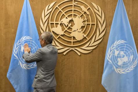 Gold UN logo on a wooden wall sits between too large blue UN flags, a dark skinned older man with greying hair wearing a grey suit stands and straightens one o the flags