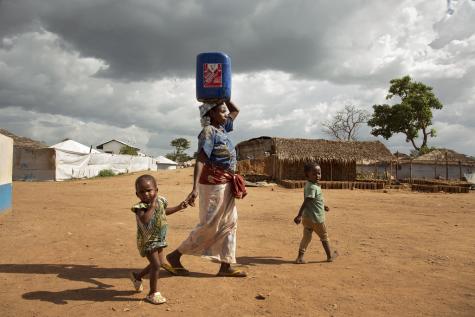 A refugee woman in Cameroon carries a heavy blue container on her head whilst walking with her two young children