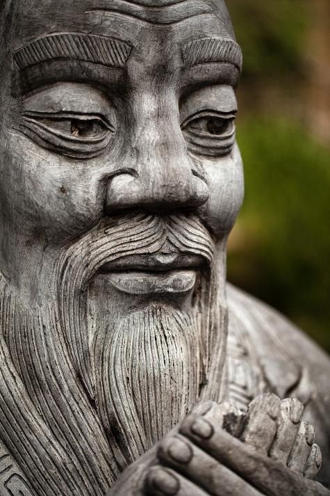 A grey statue of great philoshoper Confucius sits in a park. We mostly see his face, which looks peaceful and meditative. His hands a held together.