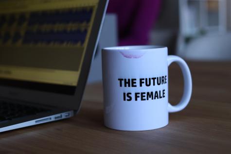 A white coffee cup with the Words ' The future is female' in black has a lipstick mark on the rim. It sits on a brown desk with a laptop in the background.